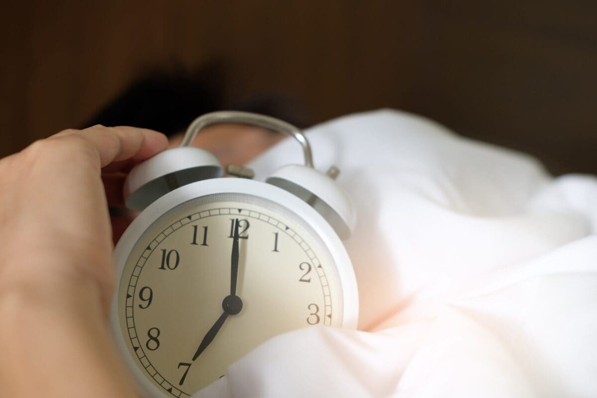 Nell'immagine si vede una mano che afferra una sveglia bianca, con le lancette che segnano il tempo. La sveglia è posizionata su un letto, coperta da un lenzuolo bianco, creando un'atmosfera tranquilla e riposante.